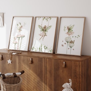 May include: Three framed watercolor prints featuring fairies. One shows a fairy on a swing, another depicts a fairy on a mushroom, and the third shows a fairy with flowers. The prints have a light wood frame and are displayed on a wooden cabinet.