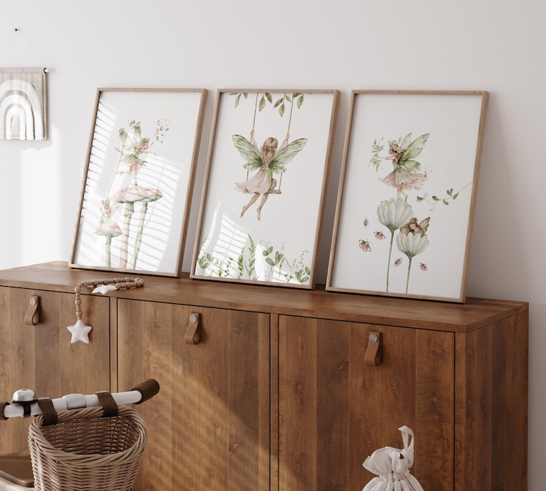 May include: Three framed watercolor prints featuring fairies. One shows a fairy on a swing, another depicts a fairy on a mushroom, and the third shows a fairy with flowers. The prints have a light wood frame and are displayed on a wooden cabinet.