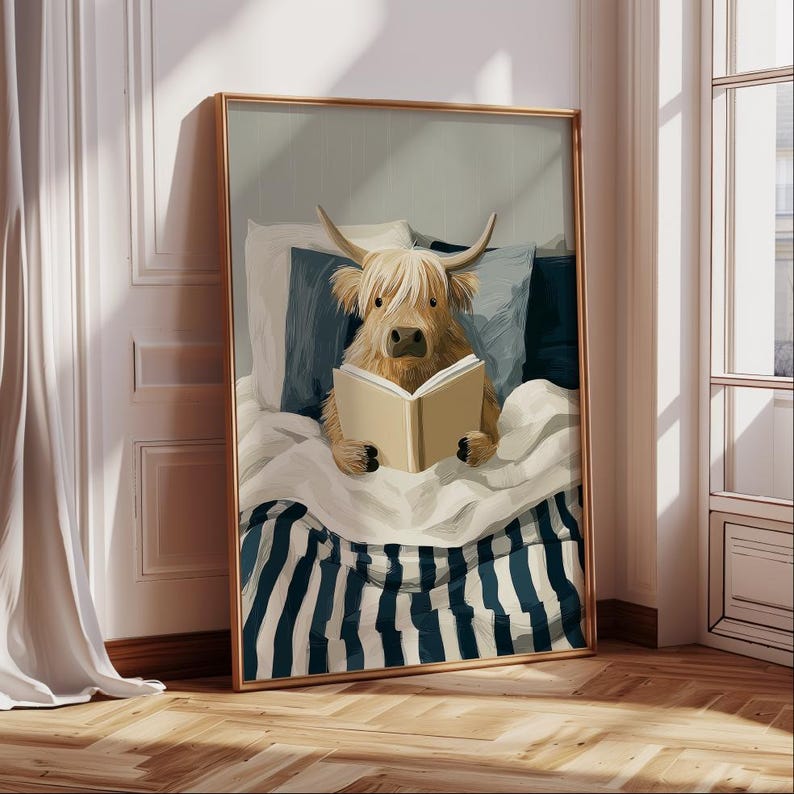 May include: A framed print showing a Highland cow reading a book in bed. The cow is light brown with long bangs and horns. The bed has blue and white striped bedding and blue pillows. The frame is gold.