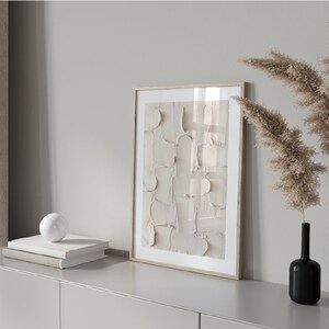 Peut inclure: Une impression d'art abstrait encadr&eacute;e aux couleurs beige et blanche est accroch&eacute;e au mur au-dessus d'une commode gris clair. Deux vases noirs avec de l'herbe de la pampa s&eacute;ch&eacute;e sont sur la commode.