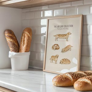Puede incluir: Una l&aacute;mina enmarcada titulada THE LAB LOAF COLLECTION Fresh from the bakery, que muestra cinco ilustraciones de perros Labrador dorados con forma de hogazas de pan, expuesta en una cocina con dos panes en un recipiente de cer&aacute;mica blanco.