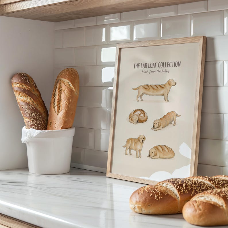 Puede incluir: Una l&aacute;mina enmarcada titulada THE LAB LOAF COLLECTION Fresh from the bakery, que muestra cinco ilustraciones de perros Labrador dorados con forma de hogazas de pan, expuesta en una cocina con dos panes en un recipiente de cer&aacute;mica blanco.