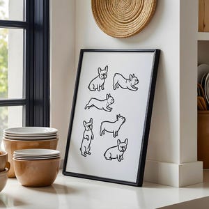 May include: Black framed art print featuring line drawings of French Bulldogs in various poses. The artwork is displayed on a white surface, leaning against a white wall. The print is in a kitchen setting, with bowls and plates nearby.
