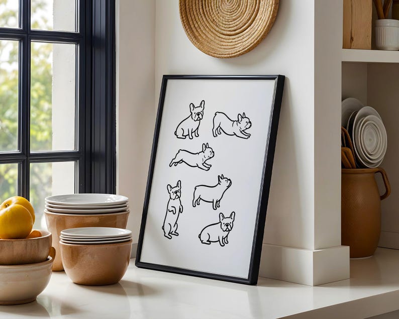 May include: Black framed art print featuring line drawings of French Bulldogs in various poses. The artwork is displayed on a white surface, leaning against a white wall. The print is in a kitchen setting, with bowls and plates nearby.