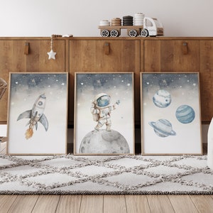 Pu&ograve; includere: Tre stampe artistiche incorniciate ad acquerello con temi spaziali. Uno mostra un razzo, un altro un astronauta sulla luna e il terzo, pianeti. L'opera d'arte ha una morbida tavolozza di colori blu, grigi e arancioni, con uno sfondo stellato.