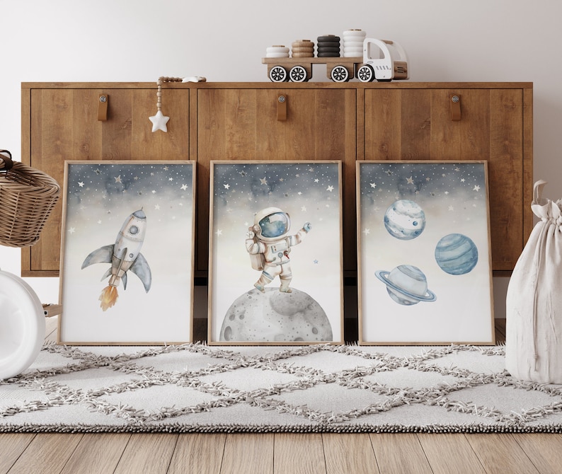 Pu&ograve; includere: Tre stampe artistiche incorniciate ad acquerello con temi spaziali. Uno mostra un razzo, un altro un astronauta sulla luna e il terzo, pianeti. L'opera d'arte ha una morbida tavolozza di colori blu, grigi e arancioni, con uno sfondo stellato.