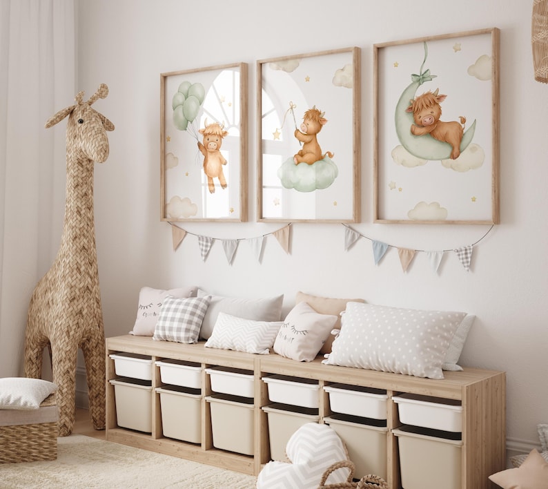 K&ouml;nnte beinhalten: Drei gerahmte Kunstdrucke f&uuml;r das Kinderzimmer mit Aquarellillustrationen eines Highland-K&auml;lbchens. Ein Kalb h&auml;lt Ballons, ein anderes sitzt auf einer Wolke, und das dritte ruht auf einem Mond. Eine gewebte Giraffe und eine Aufbewahrungsbank vervollst&auml;ndigen die Dekoration.