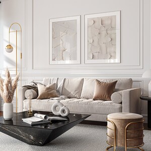 May include: A living room interior with a white sofa, two abstract art prints, a black coffee table, a gold floor lamp, and a round ottoman with a gold frame.