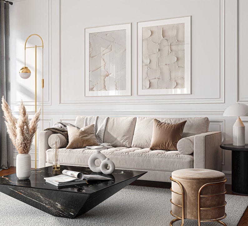 May include: A living room interior with a white sofa, two abstract art prints, a black coffee table, a gold floor lamp, and a round ottoman with a gold frame.