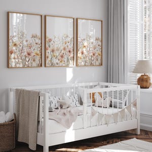 Puede incluir: Tres impresiones enmarcadas de arreglos florales de acuarela en tonos rosa, melocot&oacute;n y blanco. Las impresiones est&aacute;n colgadas en una pared blanca sobre una cuna blanca con una manta blanca y una almohada a rayas blancas y negras.