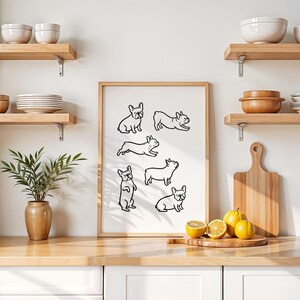 May include: A framed print featuring black line drawings of French Bulldogs in various poses. The artwork is displayed on a light-colored wall above a wooden countertop. Decorative elements include wooden shelves, bowls, and a plant in a vase.