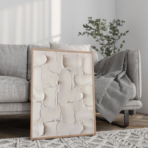 May include: A framed abstract painting with a white textured design. The painting is hanging on a wall in a living room setting with a gray couch and a throw blanket.