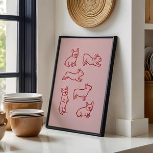 May include: A framed pink art print featuring red line drawings of French Bulldogs in various poses. The black frame leans against a white wall, near a window and a stack of white plates. A woven wall hanging is above the print.