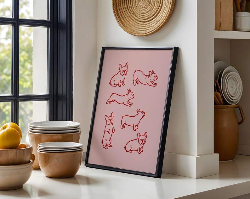 May include: A framed pink art print featuring red line drawings of French Bulldogs in various poses. The black frame leans against a white wall, near a window and a stack of white plates. A woven wall hanging is above the print.