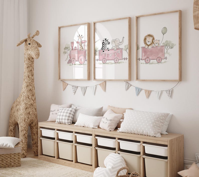 May include: Three framed wall art prints featuring watercolor illustrations of animals riding in pink train cars. The first shows a giraffe, the second a zebra and elephant, and the third a lion and cub with a green balloon, all in light wooden frames.
