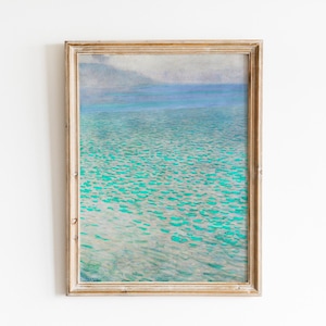 May include: A framed print of a tranquil seascape. The artwork shows turquoise water with gentle wave patterns, fading into a light blue horizon beneath a misty sky. The frame is a light, natural wood colour.