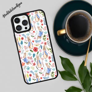 May include: Black phone case featuring a repeating pattern of watercolor bunnies in blue jackets, surrounded by colorful flowers and greenery on a white background. The case has a black border. The text "Madetobelovedyou" is in the upper left corner.