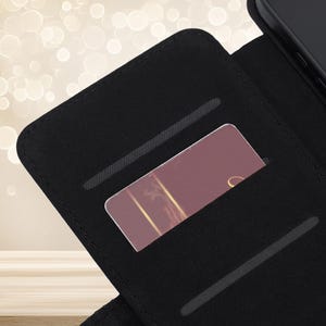 May include: A black wallet-style phone case featuring an open interior with two horizontal card slots. One slot contains a rectangular card with a dark mauve background, a thin gold vertical line, and a partial gold script letter visible on the right.