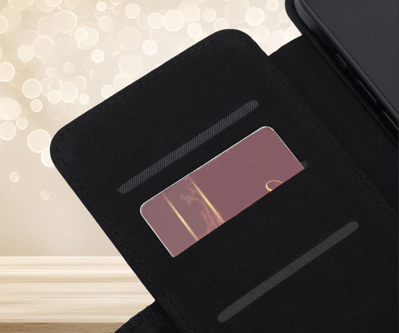 May include: A black wallet-style phone case featuring an open interior with two horizontal card slots. One slot contains a rectangular card with a dark mauve background, a thin gold vertical line, and a partial gold script letter visible on the right.