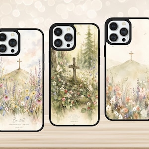 May include: Three black phone cases featuring watercolour illustrations of hills with crosses and wildflower meadows. The left and centre cases display the text "Be still, and know that I am God. Psalm 46:10" in a script and serif font combination.