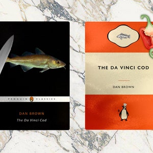 Puede incluir: Dos tablas de cortar negras y naranjas con una ilustración de pescado y el texto "The Da Vinci Cod" de Dan Brown. Una tabla tiene un cuchillo y un pescado, la otra tiene una ilustración de pingüino.