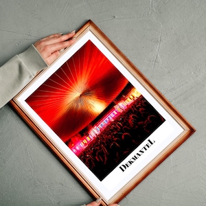 May include: A framed photograph featuring a bright red and orange light show, likely from a concert or event. The image is set in a wooden frame. The word "DEKMANTEL" is printed in white text at the bottom of the image.