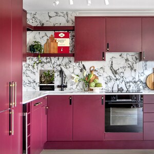 May include: A modern kitchen with burgundy cabinets and gold hardware. The backsplash is white marble with a gray veining pattern. There is a black oven and a stainless steel sink with a faucet.