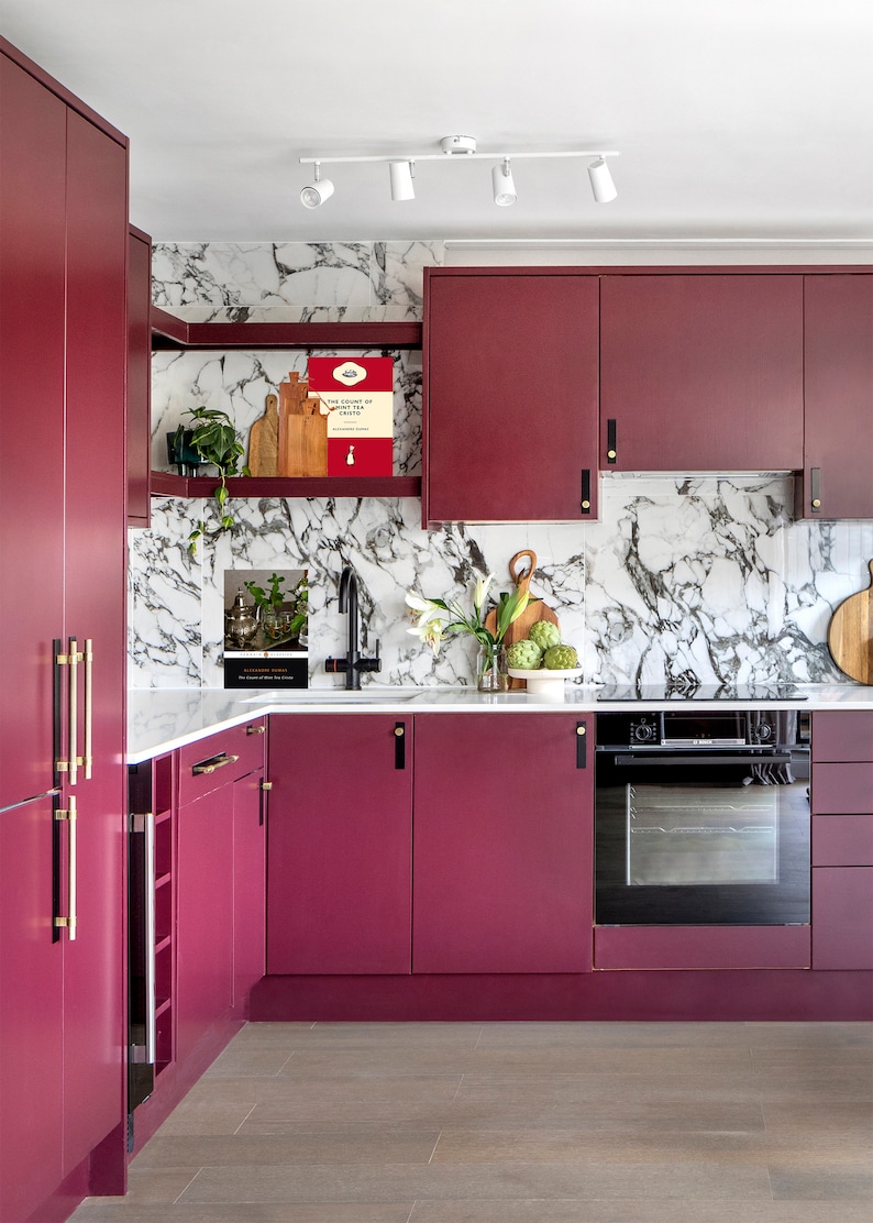 May include: A modern kitchen with burgundy cabinets and gold hardware. The backsplash is white marble with a gray veining pattern. There is a black oven and a stainless steel sink with a faucet.