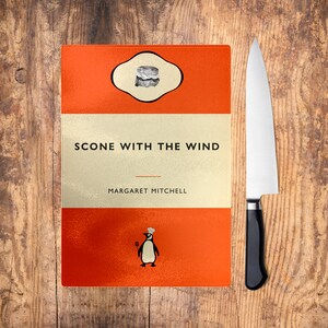 May include: A red and white cutting board with the text "Scone With The Wind" and "Margaret Mitchell" printed on it. The board features a penguin illustration and a scone icon.