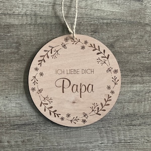 May include: A round wooden sign with a floral design and the text "Ich liebe dich Papa" in brown lettering.
