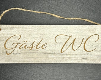 Könnte beinhalten: Rechteckiges Holzschild mit der Aufschrift "Gäste WC" in dekorativer Schrift. Das Schild ist hell mit Holzmaserung und einer Juteschnur zum Aufhängen. Der Hintergrund ist dunkel.