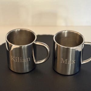 Könnte beinhalten: Zwei silberne Edelstahlbecher mit Henkeln. Der Becher links hat den Namen "Kilian" eingraviert. Der Becher rechts hat den Namen "Max" eingraviert.