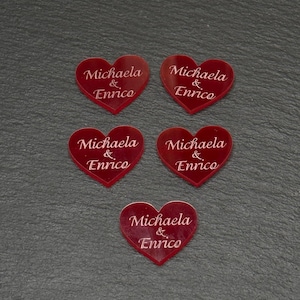 Könnte beinhalten: Fünf herzförmige, rote Acrylornamente. Jedes Herz trägt die Namen "Michaela & Enrico" in zarter, weißer Schrift eingraviert. Die Ornamente sind auf einer dunkelgrauen Oberfläche angeordnet.