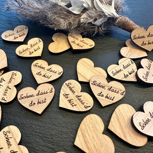 Puede incluir: Una colección de adornos de madera en forma de corazón con el texto "Schee, dass'd do bist" grabado en cada uno. Los corazones están esparcidos sobre una superficie oscura.