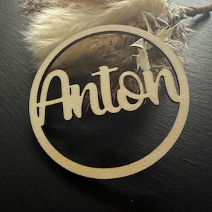 Könnte beinhalten: Ein Holzkreis mit dem ausgeschnittenen Namen Anton in der Mitte. Der Kreis befindet sich auf einem schwarzen Hintergrund.
