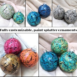 Puede incluir: Una colección de adornos navideños personalizables con salpicaduras de pintura en varios colores. Los adornos son redondos con tapas de metal plateado. Los colores incluyen verde, azul, rosa, dorado y naranja, cada uno con detalles de salpicaduras de pintura negra.