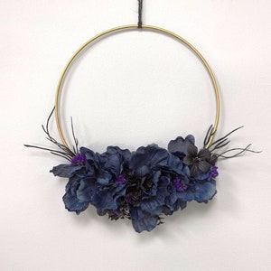 Op de afbeelding: Een gouden metalen krans versierd met donkerblauwe en paarse kunstbloemen en zwarte takken. De krans wordt opgehangen door een zwart touwtje. De bloemstuk is aan de onderkant gegroepeerd.