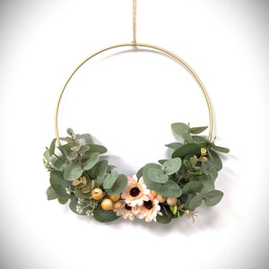 Puede incluir: Una corona circular de metal dorado adornada con hojas verdes de eucalipto, flores color melocotón y pequeñas bayas naranjas. La corona está suspendida por una cuerda de hilo natural, creando una pieza decorativa simple pero elegante.