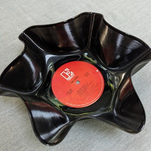 Puede incluir: Un cuenco negro de vinilo fundido con una etiqueta de disco roja y blanca en el centro. La etiqueta dice "The Cars" y "Side One Stereo". El cuenco tiene una forma ondulada única.