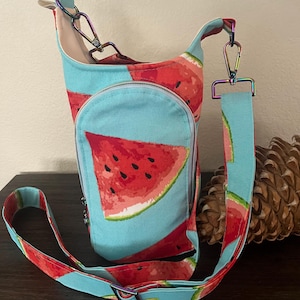 May include: A light blue crossbody bag featuring a watermelon design. The bag has a red watermelon slice pattern and an adjustable strap. The bag has a zippered pocket and iridescent hardware. Ideal for carrying essentials.