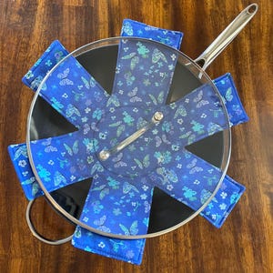 May include: A set of six blue and green pot holders with a butterfly pattern. The pot holders are arranged in a circle around a black pot with a glass lid.