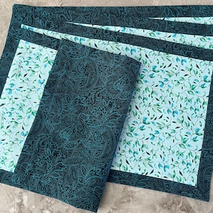 May include: Set of four teal blue placemats with a white background and a green leaf pattern. The placemats have a teal blue border.