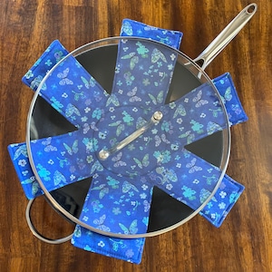 May include: A set of six blue and green pot holders with a butterfly pattern. The pot holders are arranged in a circle around a black pot with a glass lid.