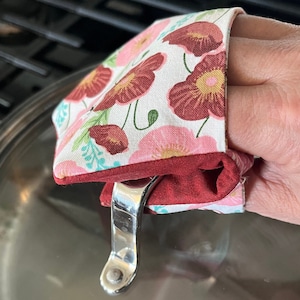 May include: A floral pot holder with a red, pink, and white floral pattern. The pot holder is made of fabric and has a red border. It is being used to hold a pot lid.