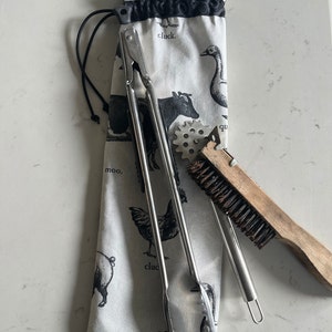 May include: A white cotton drawstring bag with black farm animal illustrations and the words "clack", "moo", and "cluck". The bag contains a pair of silver tongs, a metal grill brush, and a wooden grill brush.