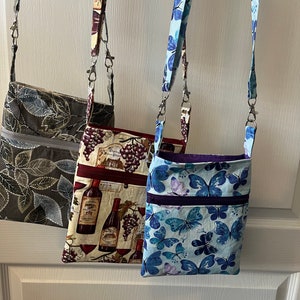 May include: Three small zippered crossbody bags with different fabric patterns. The first bag is a dark green with a leaf pattern. The second bag is a cream color with a wine bottle and grape pattern. The third bag is a light blue with a butterfly pattern.