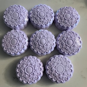 May include: Nine lavender-coloured, round bath bombs with a floral mandala design. Each bath bomb has a detailed pattern of petals and a central circular design. The bath bombs are arranged in a grid pattern.