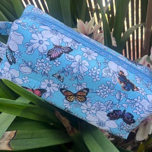 May include: A light blue zippered pouch with a floral and butterfly print. The pouch has a fabric handle and a zipper closure.