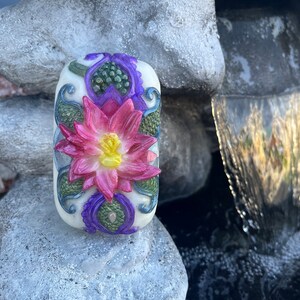 May include: A white soap bar with a pink and purple floral design. The soap features a lotus flower with green leaves and a paisley pattern.