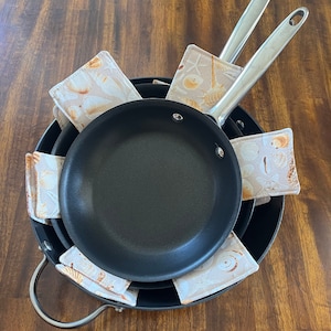 May include: A set of black non-stick frying pans nested together with beige fabric pan protectors featuring a seashell and starfish print placed between each pan to prevent scratching and damage during storage.